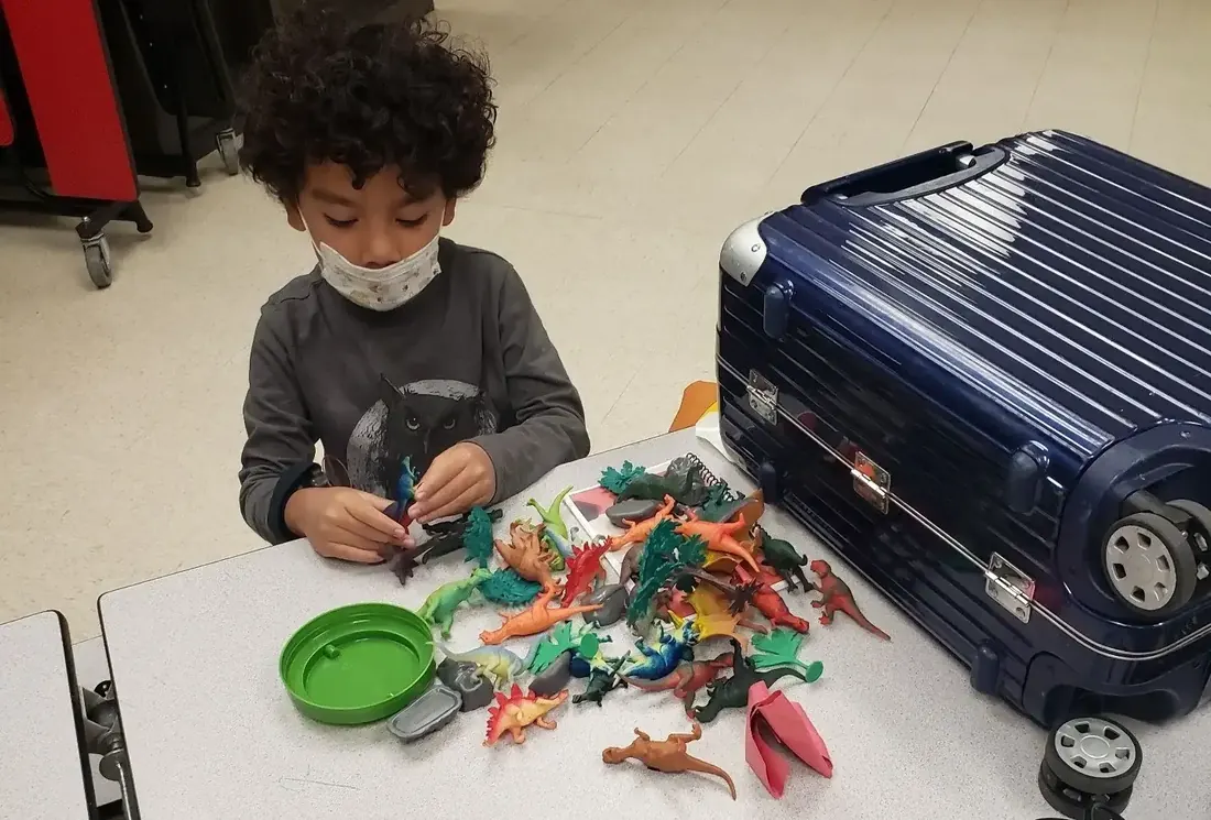 A boy plays with a pile of toy dinosaurs at a table; the story book that came with them goes unread