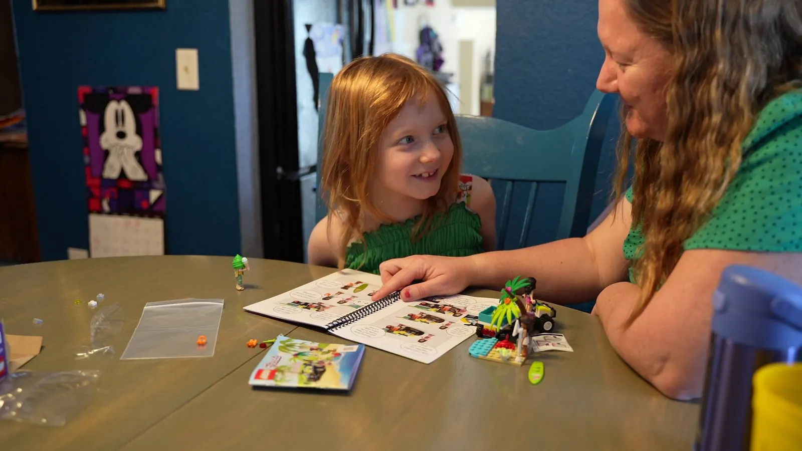 A girl grins at an adult while they read a LEGO story book together at a kitchen table