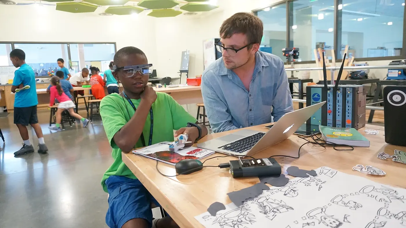 A smiling boy wearing eye-tracking glasses reads on a laptop while a researcher sits beside him